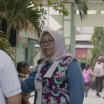 Seorang perempuan berbincang di lingkungan sekolah dasar di Surabaya, dengan siswa berseragam di latar belakang.