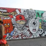 Dinding panjang bergambar mural warna-warni di Surabaya, dengan seorang pejalan kaki melintas di depannya.