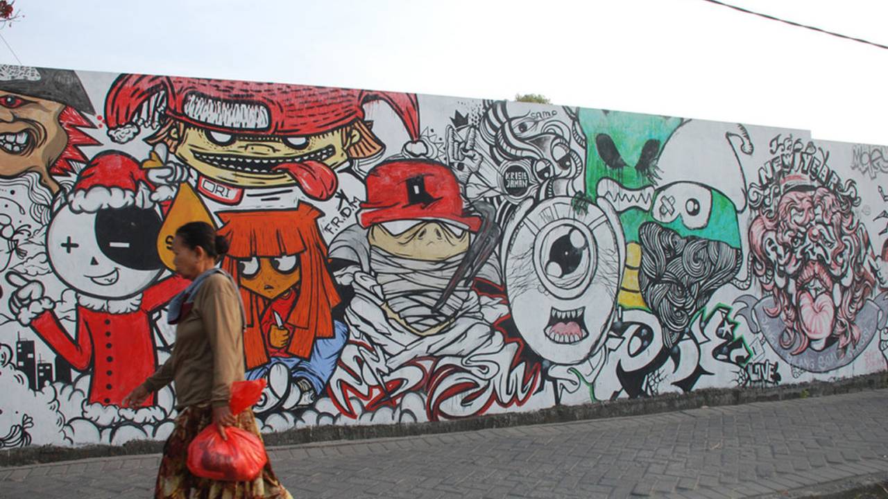 Dinding panjang bergambar mural warna-warni di Surabaya, dengan seorang pejalan kaki melintas di depannya.